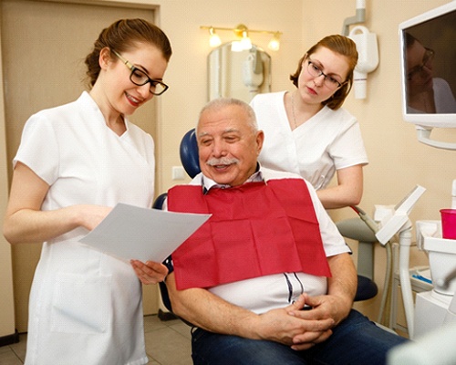 : An older man sitting in a dentist’s chair while dental workers speak with him